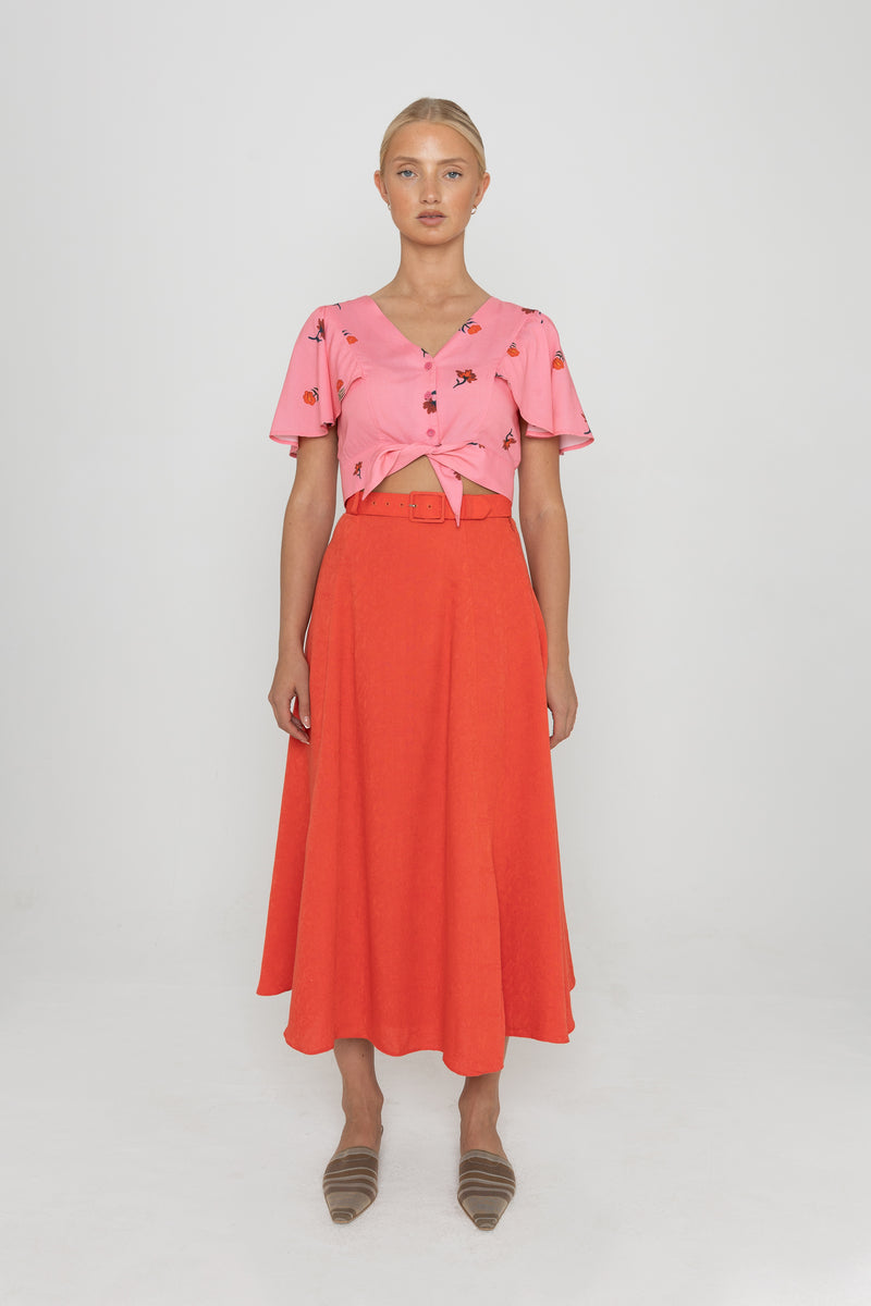 Woman wearing a Deidei Beatrix top with  a pink floral rose print and a red skirt on a white background