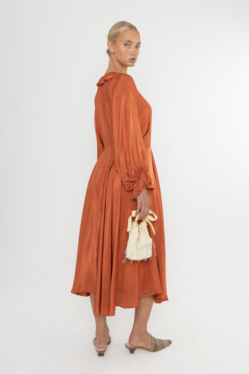 Woman wearing a rust-colored satin dress with long sleeves  and a ruffle collar with brown mules and a yellow bag on a white background
