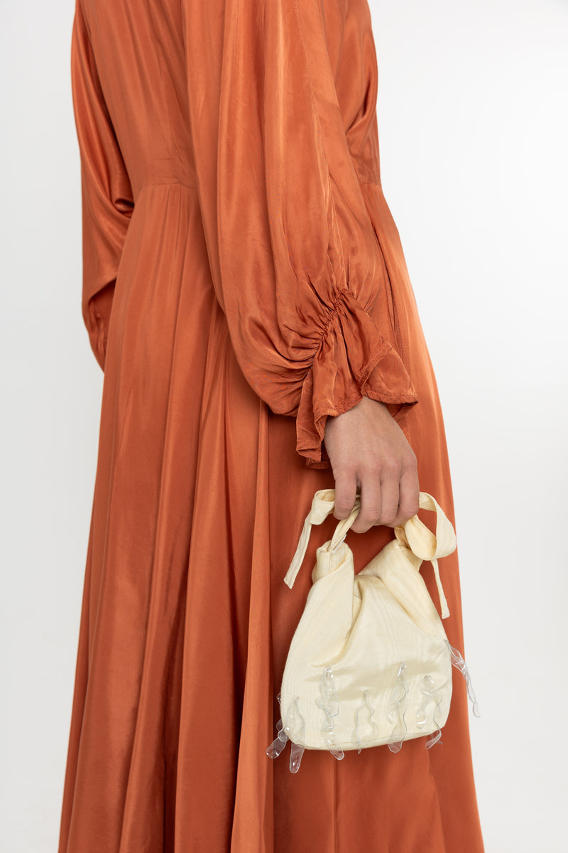 Woman wearing a rust-colored satin dress with long sleeves  and a ruffle collar with a yellow bag on a white background