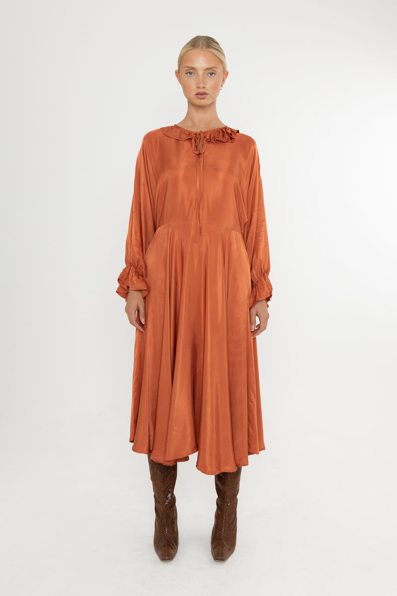 Woman wearing a rust-colored satin dress with long sleeves  and a ruffle collar with brown boots on a white background