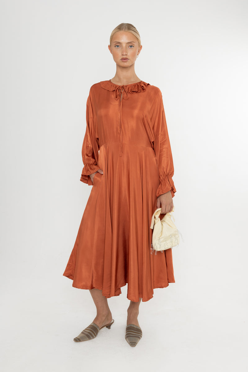 Woman wearing a rust-colored satin dress with long sleeves  and a ruffle collar with brown mules and a yellow bag on a white background