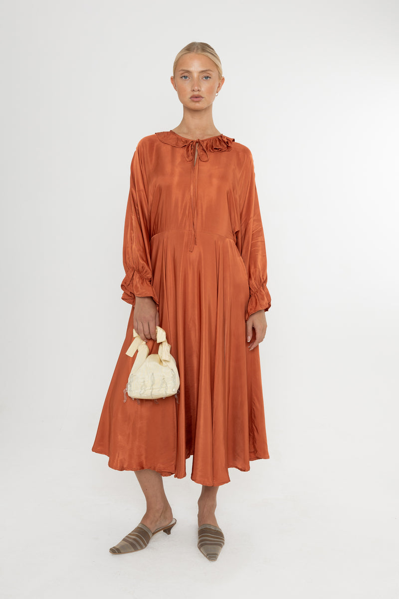 Woman wearing a rust-colored satin dress with long sleeves  and a ruffle collar with brown mules and a yellow bag on a white background