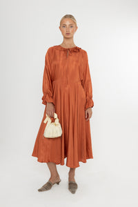 Woman wearing a rust-colored satin dress with long sleeves  and a ruffle collar with brown mules and a yellow bag on a white background