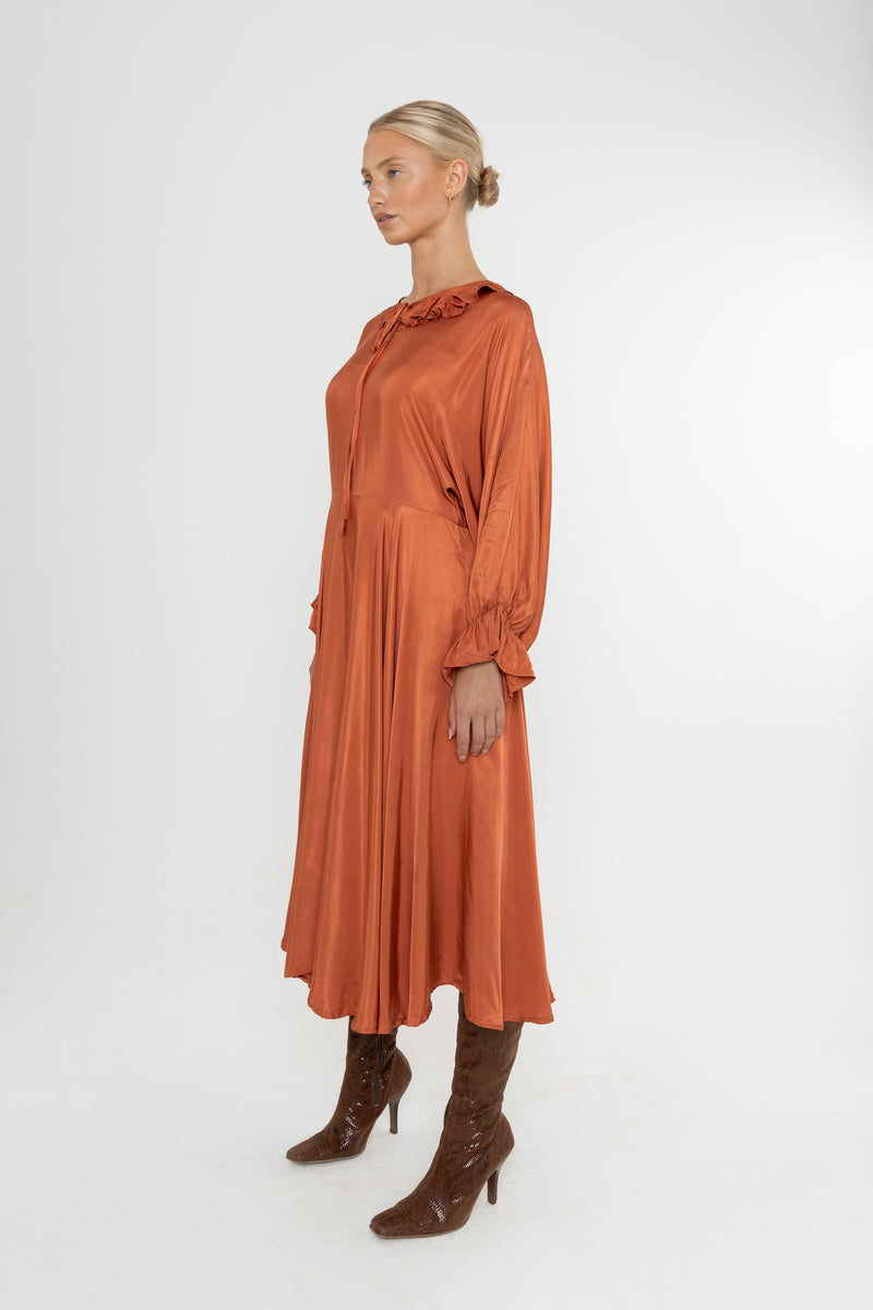 Woman wearing a rust-colored satin dress with long sleeves  and a ruffle collar with brown boots on a white background