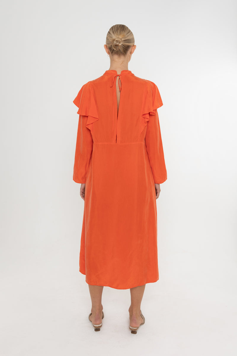 Woman wearing an orange vermillion red dress with ruffled long sleeves and mules on a white background