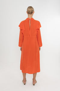 Woman wearing an orange vermillion red dress with ruffled long sleeves and mules on a white background