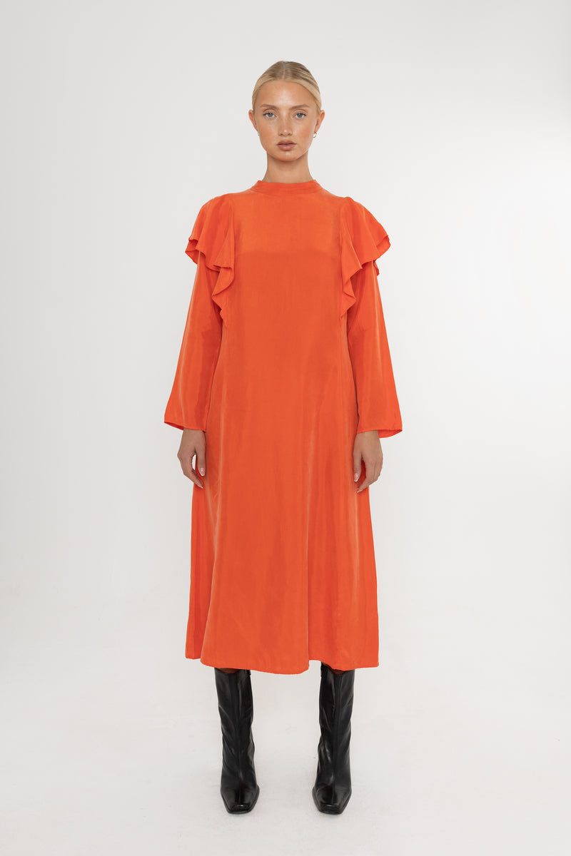 Woman wearing an orange vermillion red dress with ruffled long sleeves and black boots on a white background