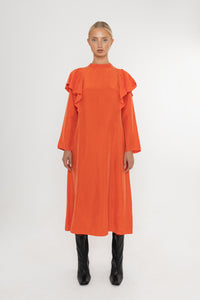 Woman wearing an orange vermillion red dress with ruffled long sleeves and black boots on a white background