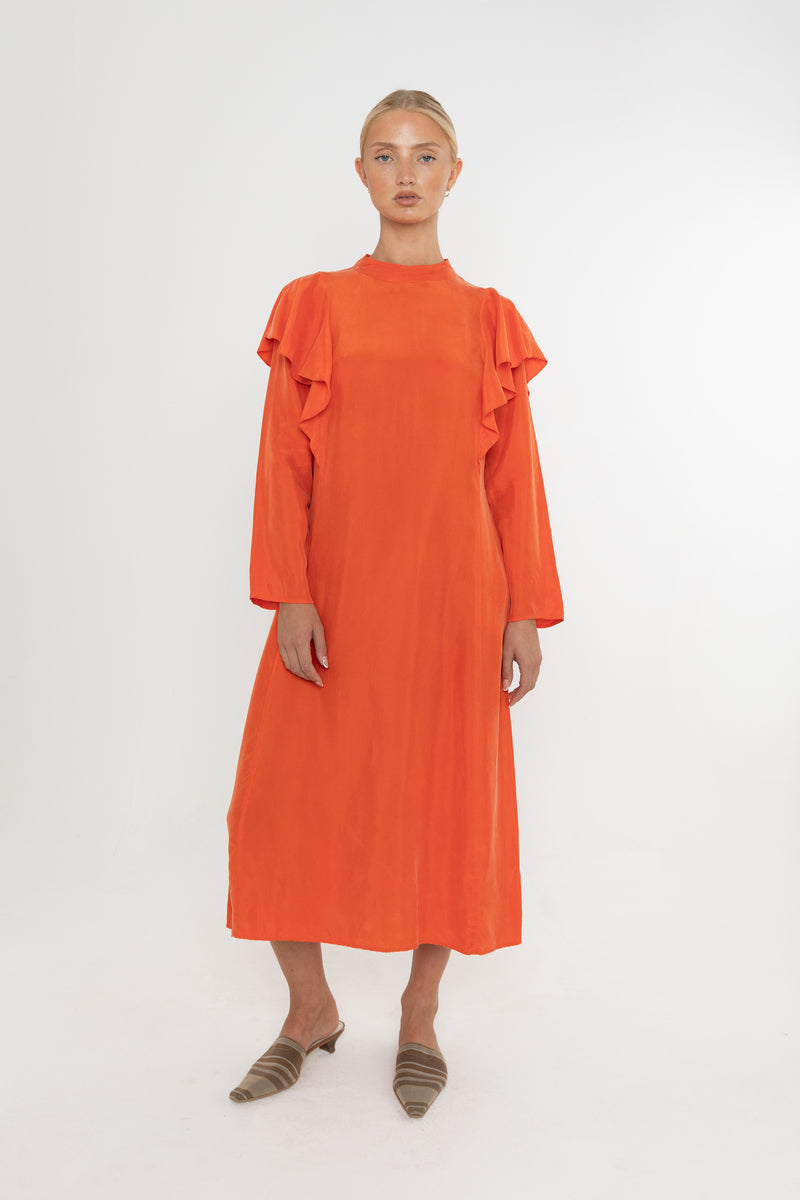 Woman wearing an orange vermillion red dress with ruffled long sleeves and mules on a white background