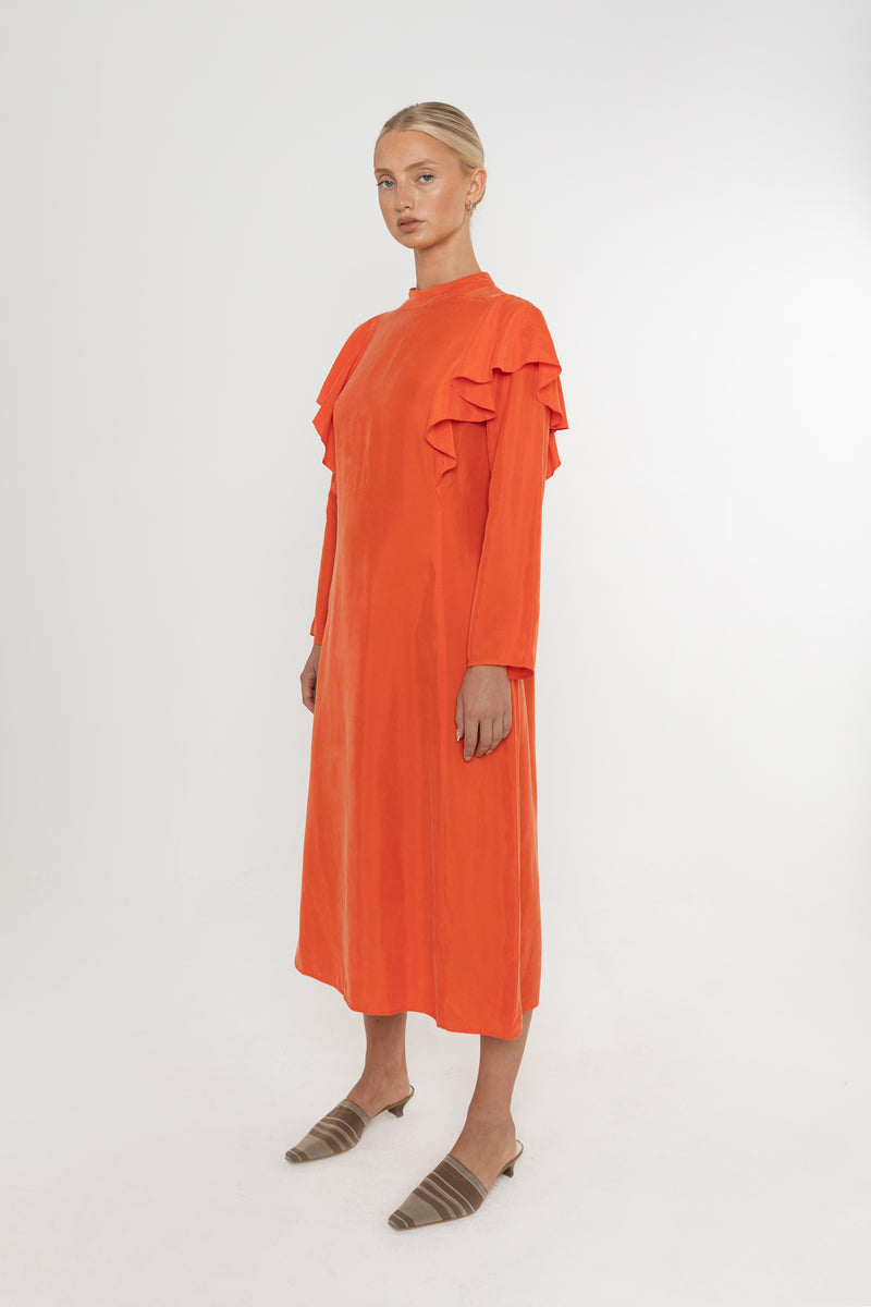 Woman wearing an orange vermillion red dress with ruffled long sleeves and brown mules on a white background