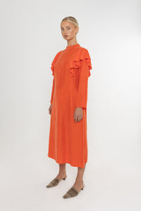 Woman wearing an orange vermillion red dress with ruffled long sleeves and brown mules on a white background