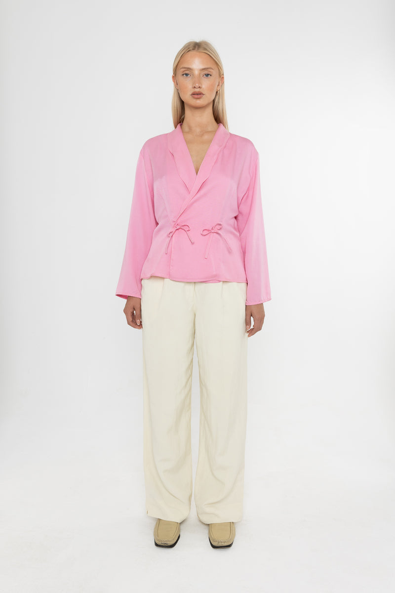 Woman wearing a Deidei Malala pink blazer jacket and white trousers holding a matching pink bag on a white background