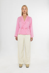 Woman wearing a Deidei Malala pink blazer jacket and white trousers holding a matching pink bag on a white background