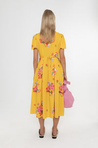 Woman wearing a Deidei Maya midi dress in a yellow floral print with a pink bag on a white background