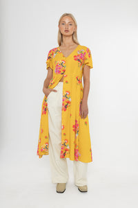 Woman wearing a Deidei Maya midi dress in a yellow floral print with over trousers on a white background