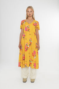Woman wearing a Deidei Maya midi dress in a yellow floral print with over trousers on a white background