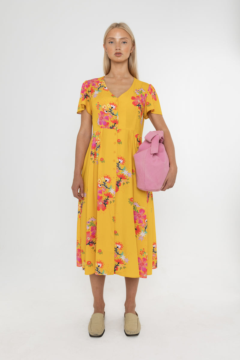 Woman wearing a Deidei Maya midi dress in a yellow floral print holding a pink bag on a white background