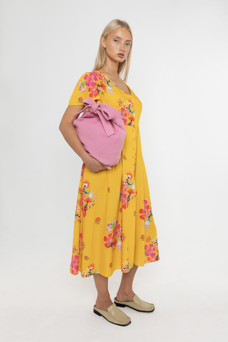 Woman wearing a Deidei Maya midi dress in a yellow floral print holding a pink bag on a white background