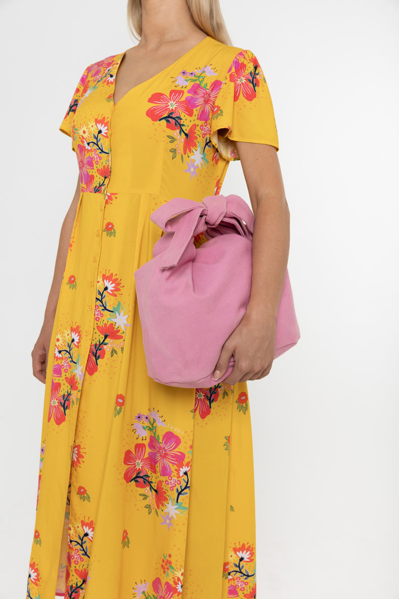 Woman wearing a Deidei Maya midi dress in a yellow floral print holding a pink bag on a white background