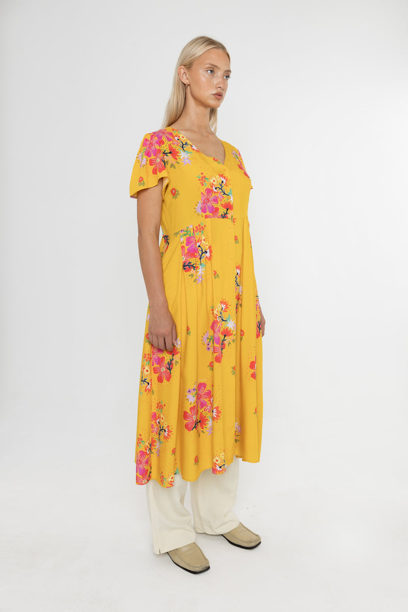 Woman wearing a Deidei Maya midi dress in a yellow floral print with over trousers on a white background