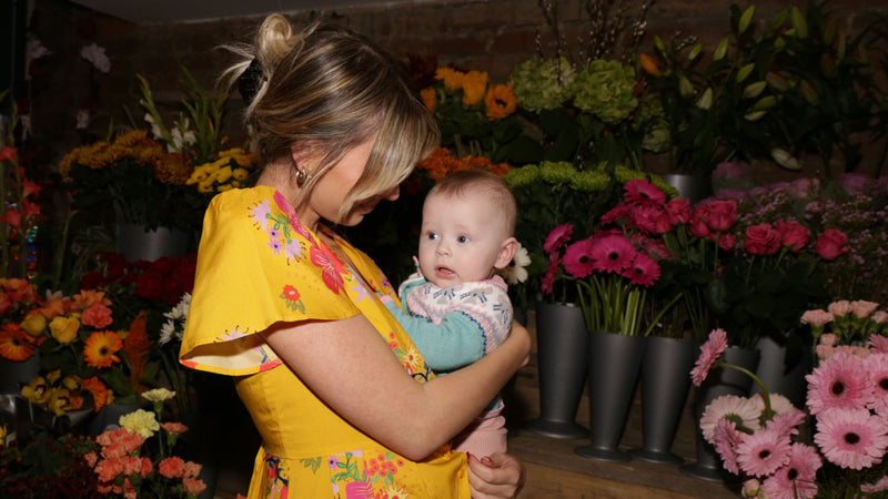Model wears a deidei maya midi dress in yellow whilst holding her baby in a florists