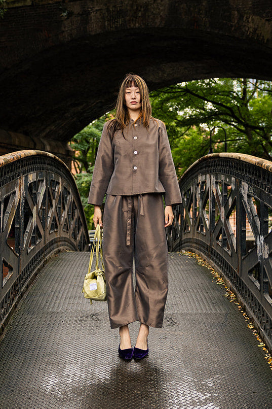 A woman stands on a wet arched bridge under an overpass, wearing a matching taupe-brown two-piece outfit with a cropped jacket and wide-leg pants, holding a small yellow bag.