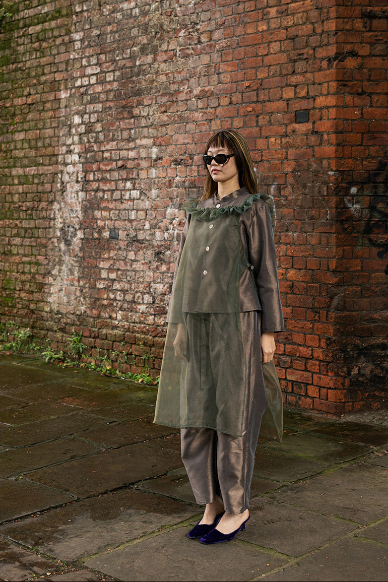 A woman wearing a muted olive green two-piece outfit with a ruffled collar and matching pants, standing against a weathered red-brown brick wall.