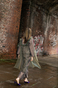 A woman wearing a gray-green ruffled coat and matching trousers walks away in an urban brick tunnel setting.