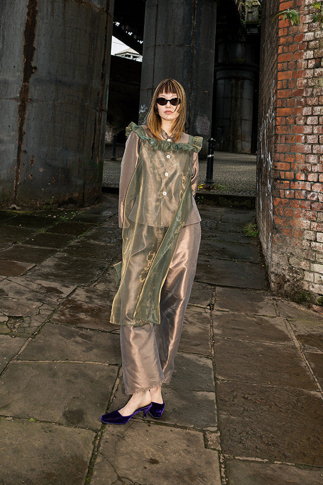 A woman wearing a sheer, metallic sage green cape-style top and matching pants stands on a stone walkway between large industrial metal columns and a brick wall.