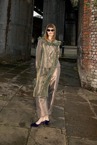 A woman wearing a sheer, metallic sage green cape-style top and matching pants stands on a stone walkway between large industrial metal columns and a brick wall.