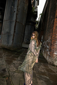 A woman wearing a shiny taupe satin robe and matching pants stands in an industrial alleyway with large metal pipes and brick walls.