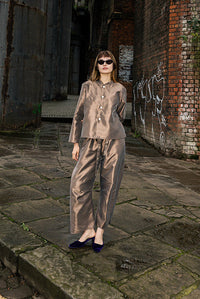 A woman wearing a matching beige two-piece suit stands on a stone walkway between large industrial brick and metal structures.