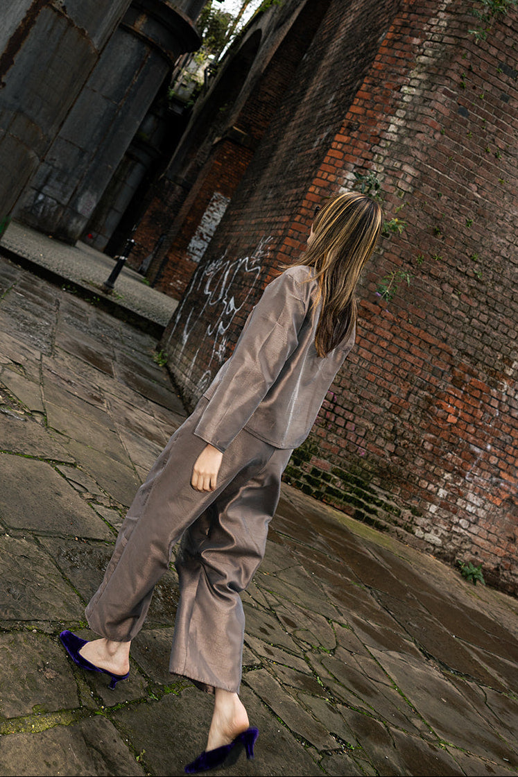 A woman wearing a loose taupe-gray jumpsuit and dark blue pointed-toe heels walks barefoot on a stone pathway under a large industrial structure and brick wall.