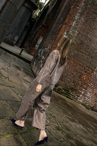 A woman wearing a loose taupe-gray jumpsuit and dark blue pointed-toe heels walks barefoot on a stone pathway under a large industrial structure and brick wall.