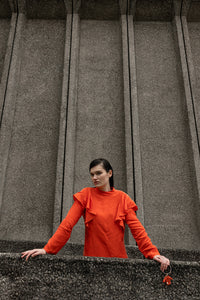 A woman wearing a bright orange long-sleeve top with ruffle details on the shoulders, standing in front of a large textured gray stone wall.