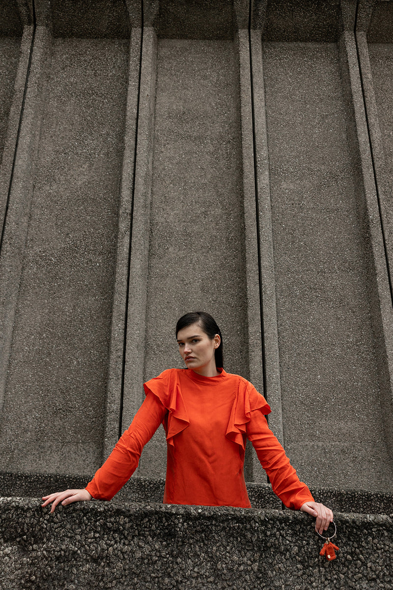 A woman wearing a bright orange long-sleeve top with ruffle details on the shoulders, standing in front of a large textured gray stone wall.