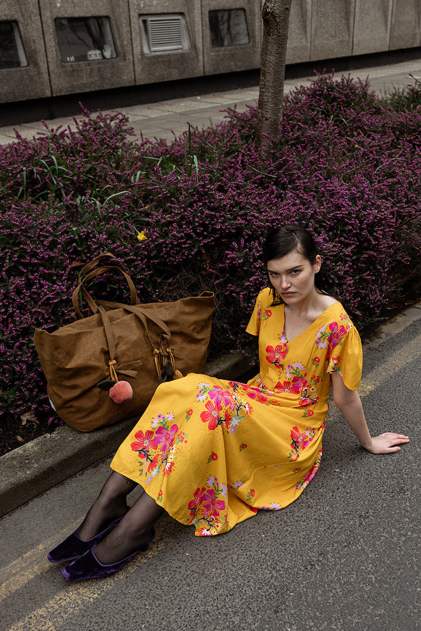 A woman sits on a paved street next to a curb, wearing a bright yellow floral midi dress and holding a brown tote bag. The background features purple foliage, a concrete building, and a tree.