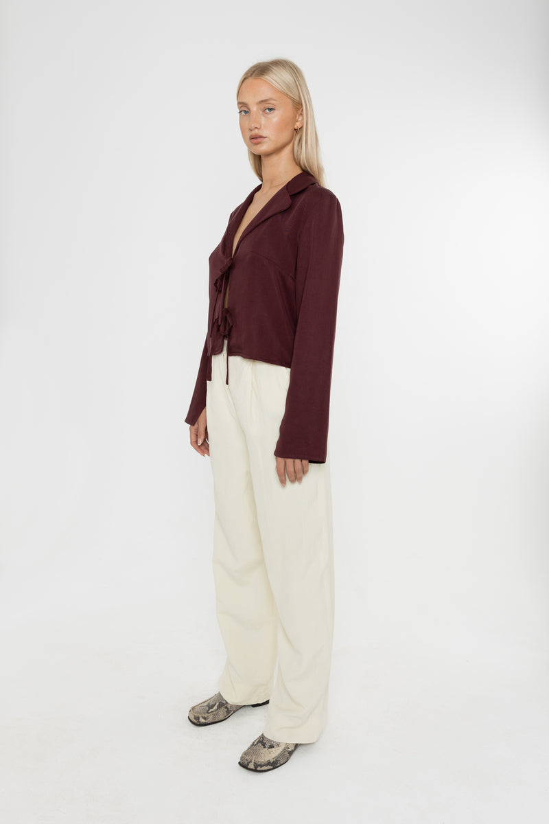 Woman wearing a burgundy brown jacket and white trousers with snake print loafers on a white background