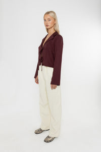 Woman wearing a burgundy brown jacket and white trousers with snake print loafers on a white background