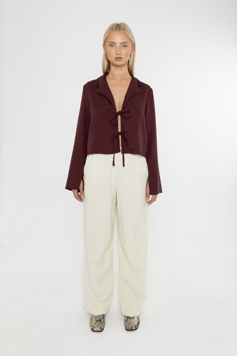Woman wearing a burgundy brown jacket and white trousers with snake print loafers on a white background