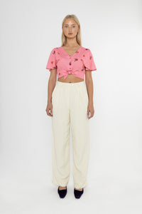 Woman wearing a Deidei Beatrix top with  a pink floral rose print and white trousers on a white background