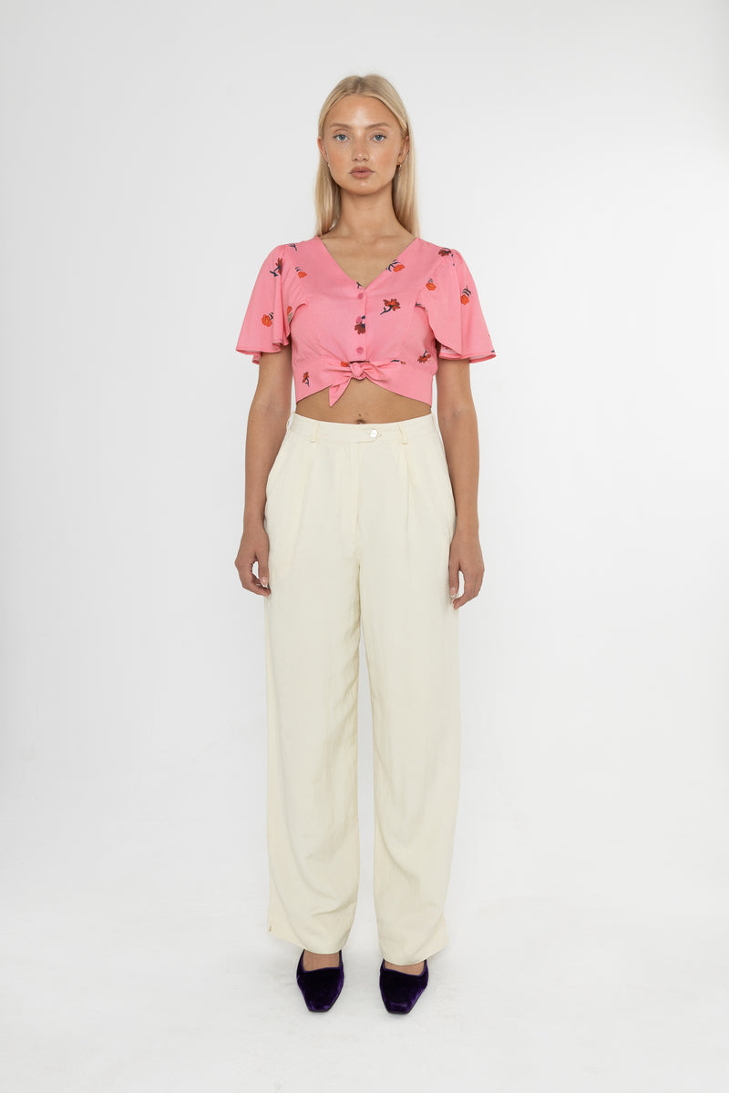 Woman wearing a Deidei Beatrix top with  a pink floral rose print and white trousers on a white background