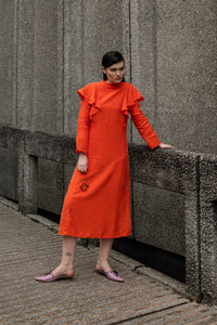 A woman wearing a bright orange long-sleeve midi dress with ruffled shoulders and pink pointed flats stands on a concrete walkway beside a textured gray concrete wall.
