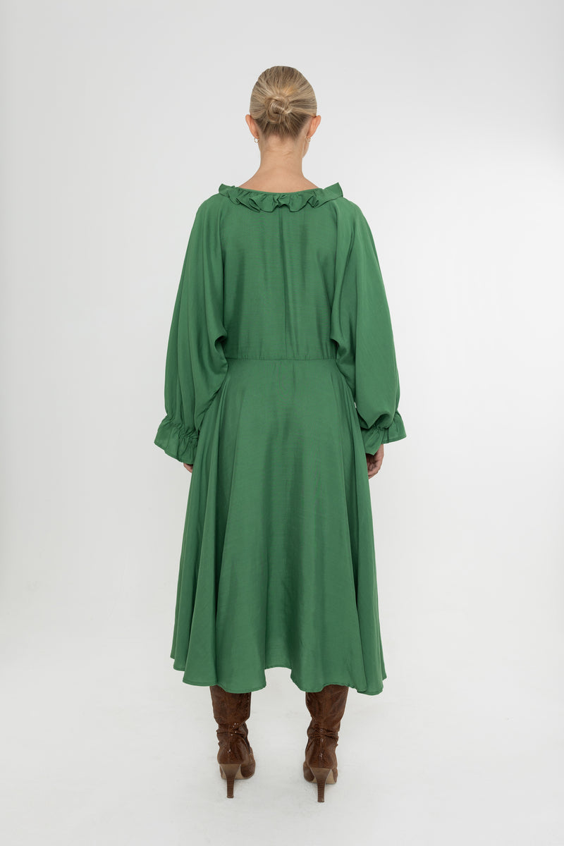 Woman wearing a green Deidei Florence dress with long sleeves and a ruffle collar with brown boots on a white background