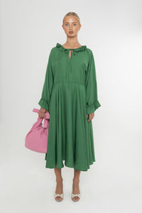 Woman wearing a green Deidei Florence dress with long sleeves and a ruffle collar with a pink bag and white shoes on a white background