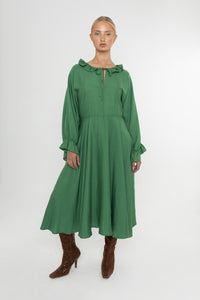 Woman wearing a green Deidei Florence dress with long sleeves and a ruffle collar with brown boots on a white background