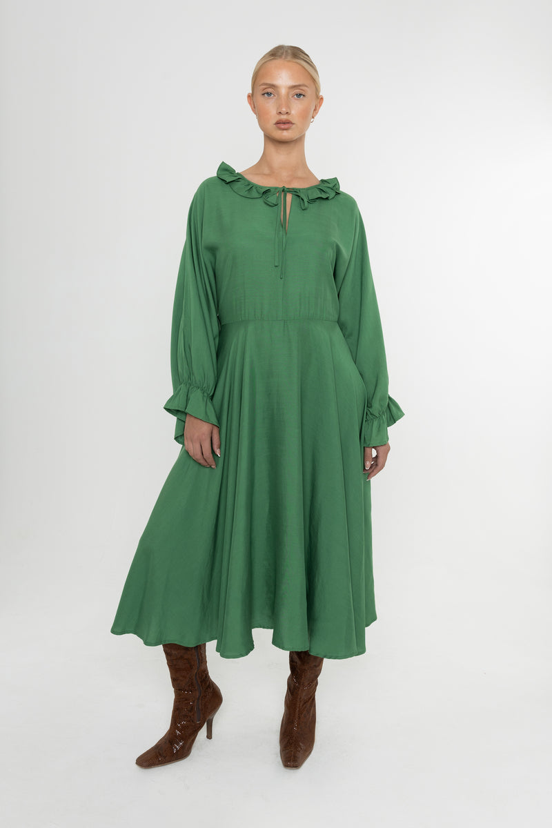 Woman wearing a green Deidei Florence dress with long sleeves and a ruffle collar with brown boots on a white background