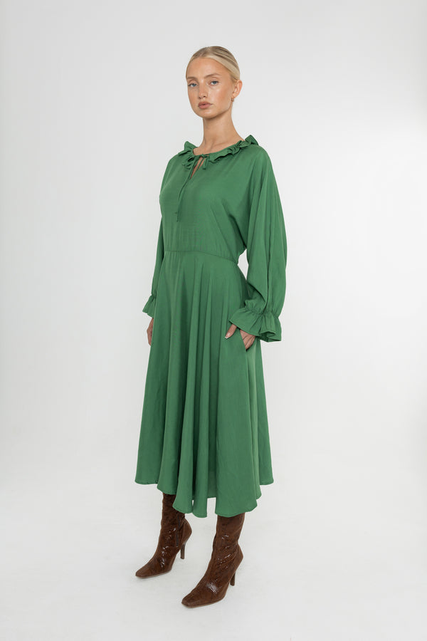 Woman wearing a green Deidei Florence dress with long sleeves and a ruffle collar with brown boots on a white background