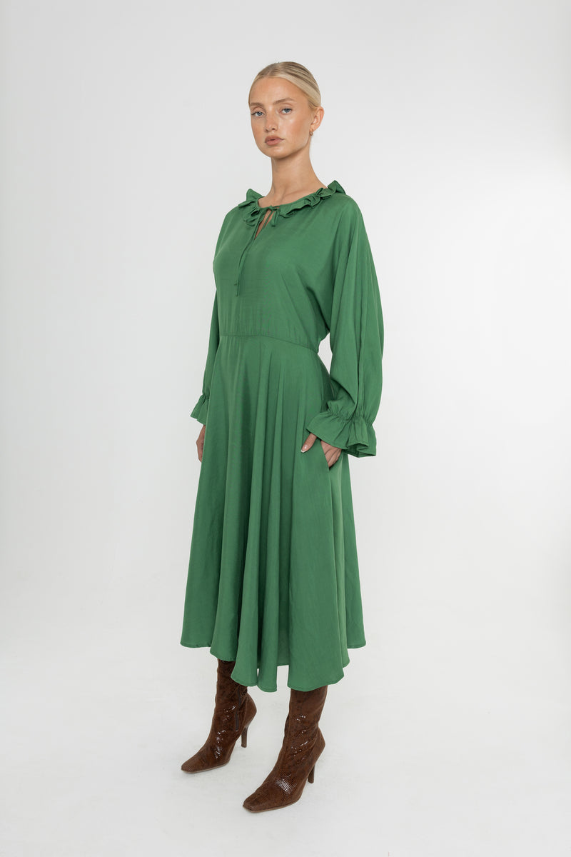 Woman wearing a green Deidei Florence dress with long sleeves and a ruffle collar with brown boots on a white background