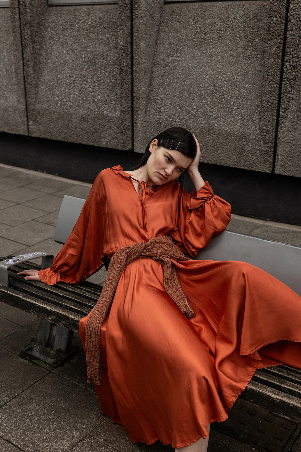 Woman in an orange dress sitting on a bench against a textured wall.